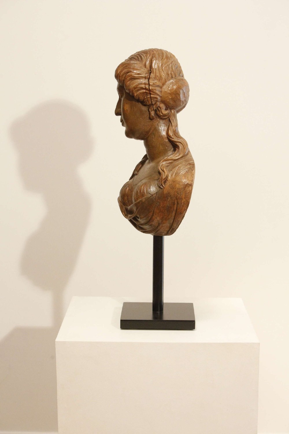 An antique carved wood bust of a maiden with styled wavy hair and draped clothing, mounted on a black metal display stand. View XI