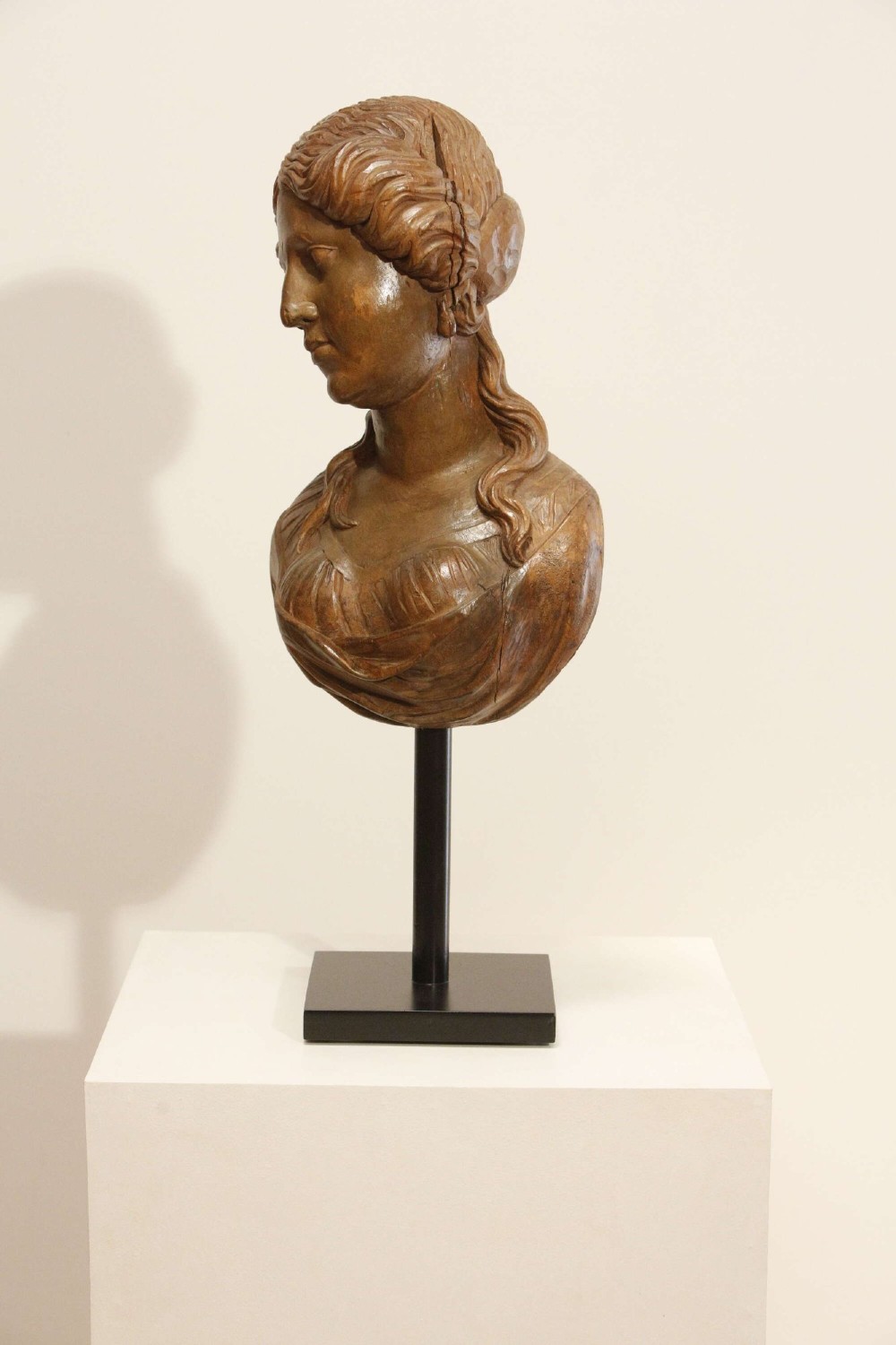 An antique carved wood bust of a maiden with styled wavy hair and draped clothing, mounted on a black metal display stand. View XII
