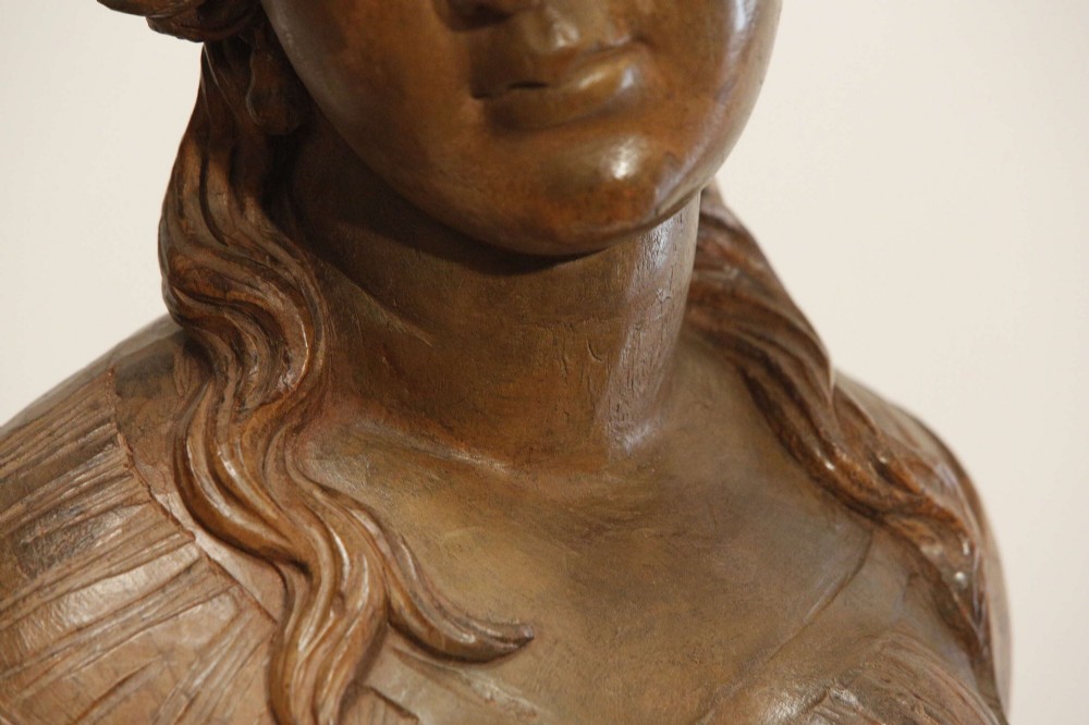 An antique carved wood bust of a maiden with styled wavy hair and draped clothing, mounted on a black metal display stand. View XIII