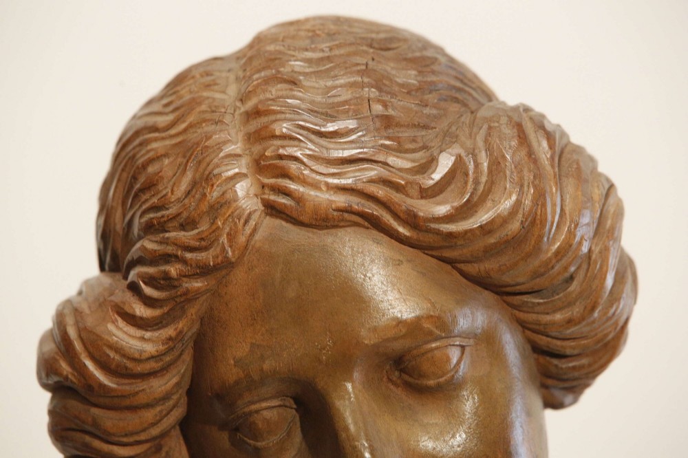 An antique carved wood bust of a maiden with styled wavy hair and draped clothing, mounted on a black metal display stand. View XIV