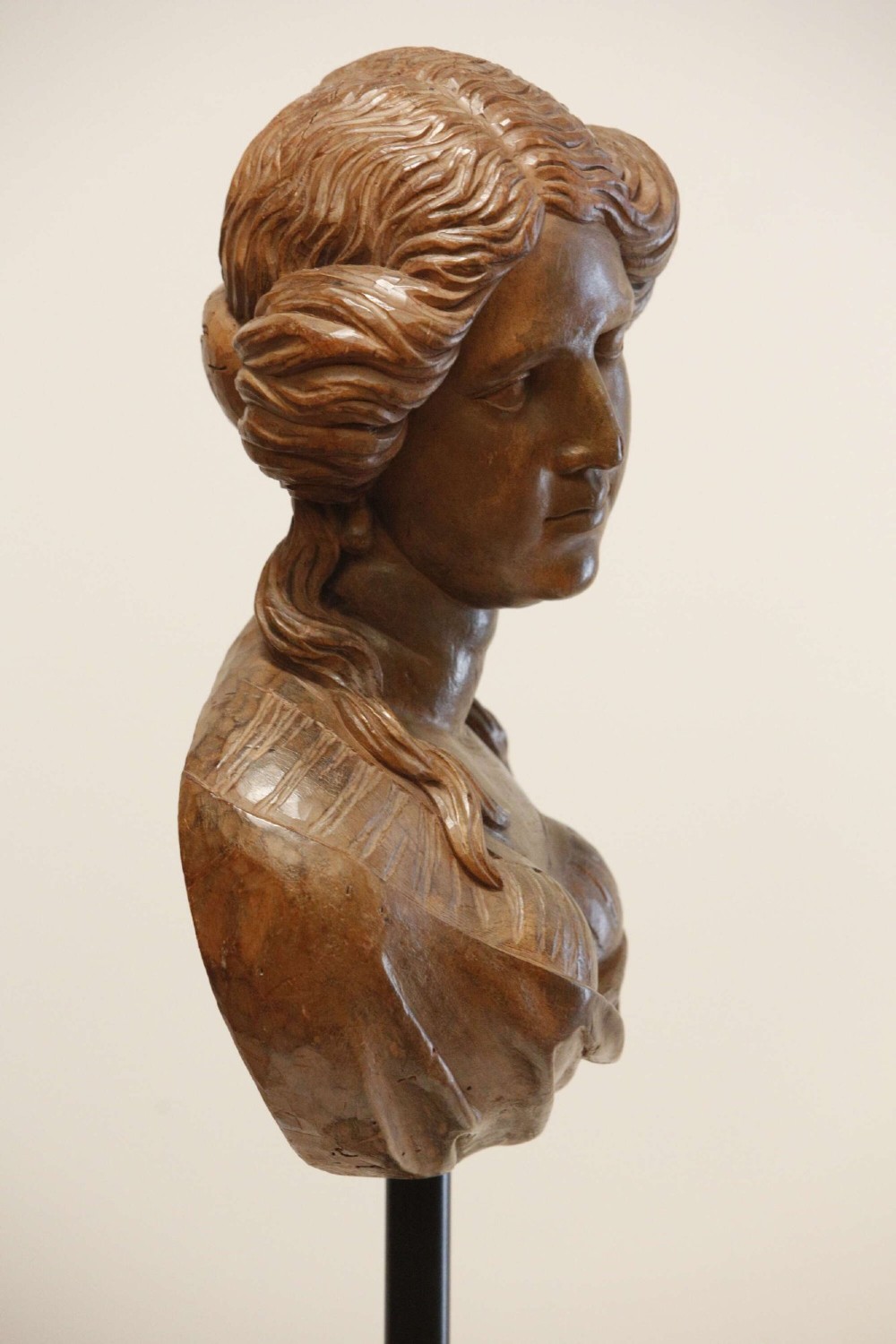 An antique carved wood bust of a maiden with styled wavy hair and draped clothing, mounted on a black metal display stand. View XV
