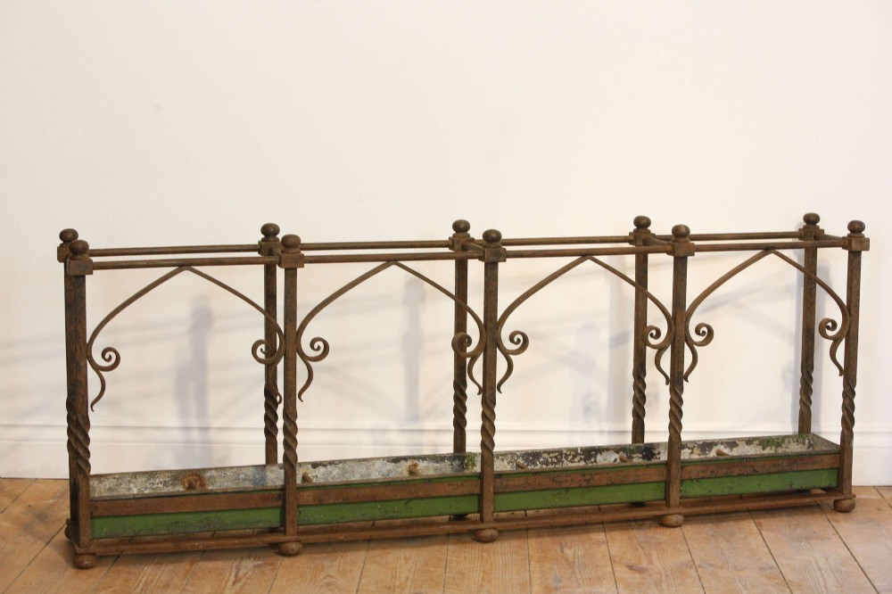 A rectangular Victorian Gothic Revival wrought iron umbrella stand featuring pointed tracery arches, twisted corner posts with ball finials, and a removable green metal drip tray. View II