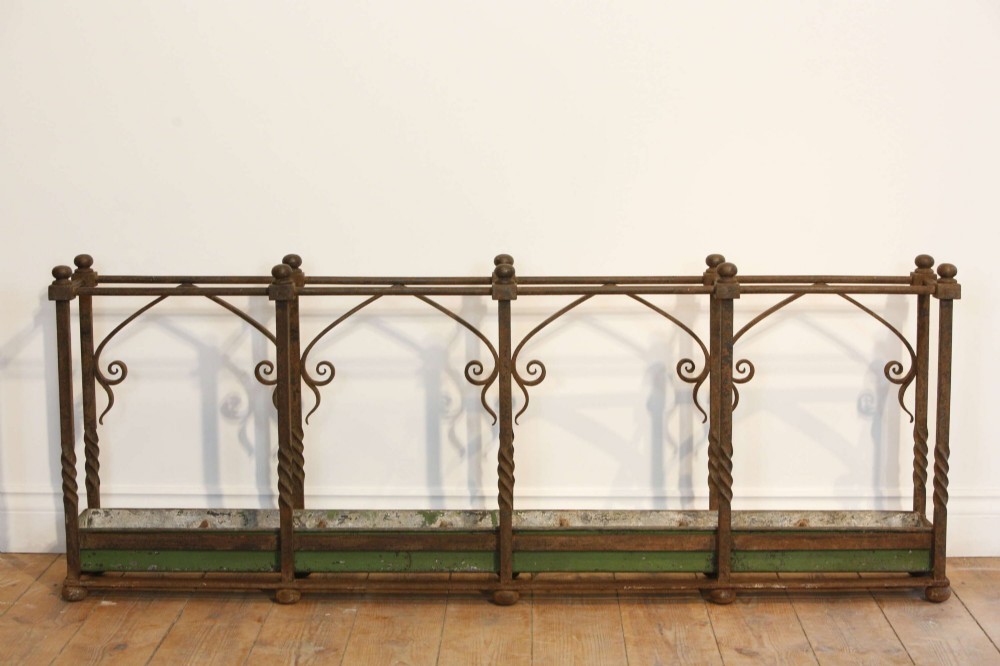A rectangular Victorian Gothic Revival wrought iron umbrella stand featuring pointed tracery arches, twisted corner posts with ball finials, and a removable green metal drip tray. View V