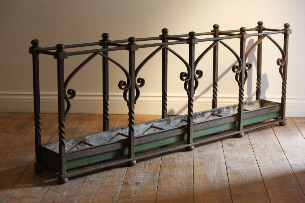 A rectangular Victorian Gothic Revival wrought iron umbrella stand featuring pointed tracery arches, twisted corner posts with ball finials, and a removable green metal drip tray. View VI
