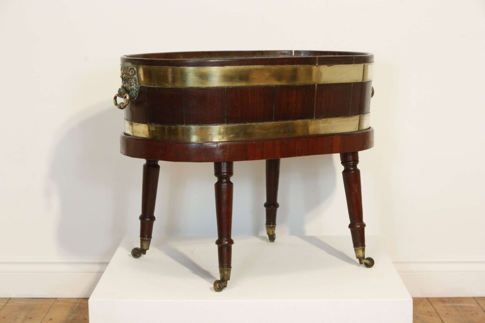 An oval mahogany wine cooler featuring twin brass binding, lion mask ring handles, tapered turned legs with brass castors, and a zinc liner. View II