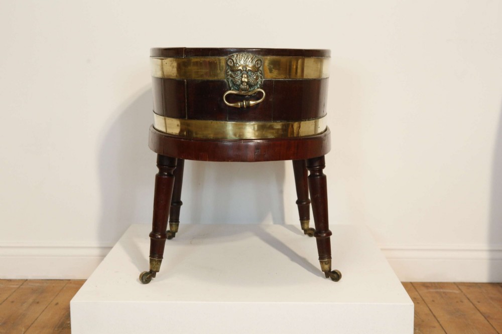 An oval mahogany wine cooler featuring twin brass binding, lion mask ring handles, tapered turned legs with brass castors, and a zinc liner. View III