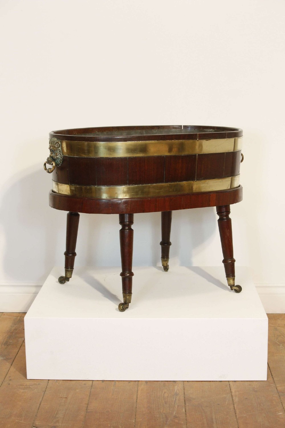 An oval mahogany wine cooler featuring twin brass binding, lion mask ring handles, tapered turned legs with brass castors, and a zinc liner. View IV