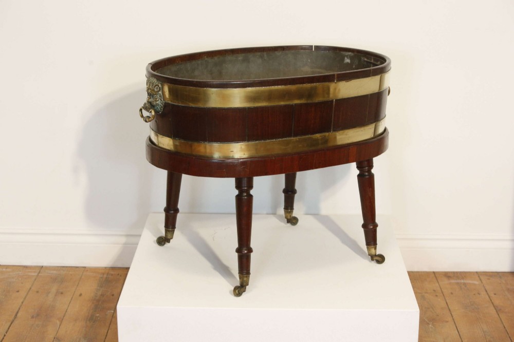 An oval mahogany wine cooler featuring twin brass binding, lion mask ring handles, tapered turned legs with brass castors, and a zinc liner. View V