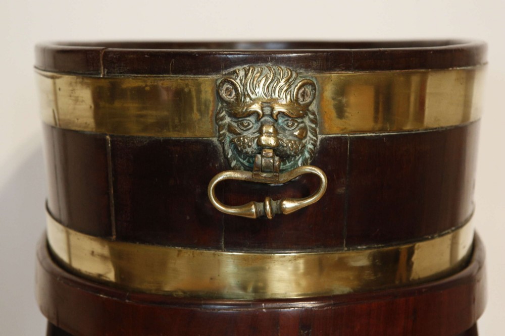 An oval mahogany wine cooler featuring twin brass binding, lion mask ring handles, tapered turned legs with brass castors, and a zinc liner. View VI