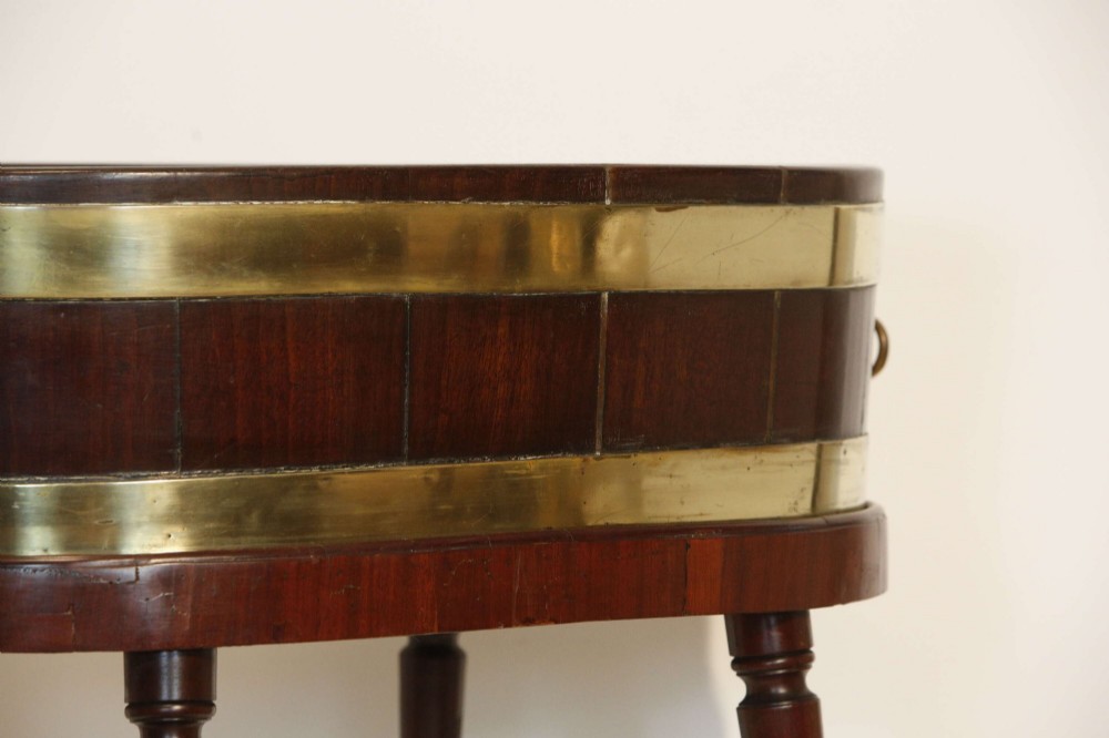 An oval mahogany wine cooler featuring twin brass binding, lion mask ring handles, tapered turned legs with brass castors, and a zinc liner. View VII