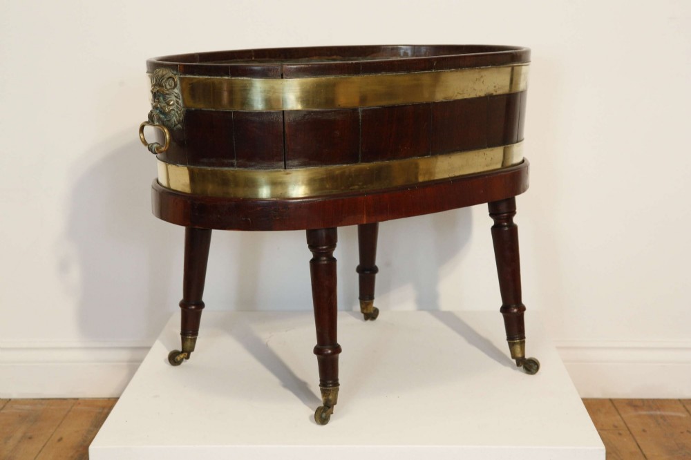 An oval mahogany wine cooler featuring twin brass binding, lion mask ring handles, tapered turned legs with brass castors, and a zinc liner. View VIII