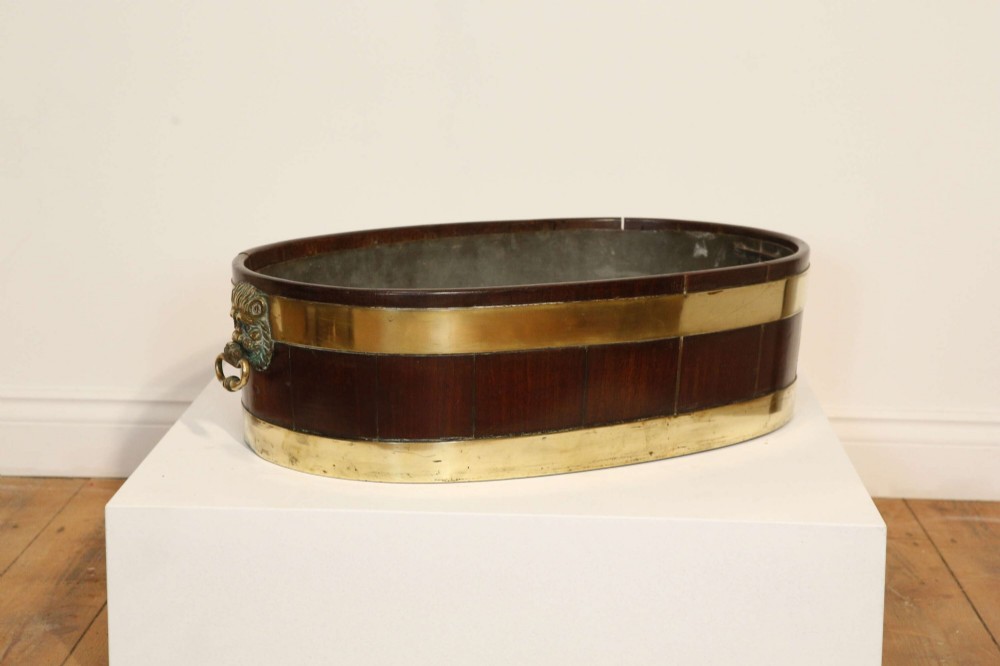 An oval mahogany wine cooler featuring twin brass binding, lion mask ring handles, tapered turned legs with brass castors, and a zinc liner. View X