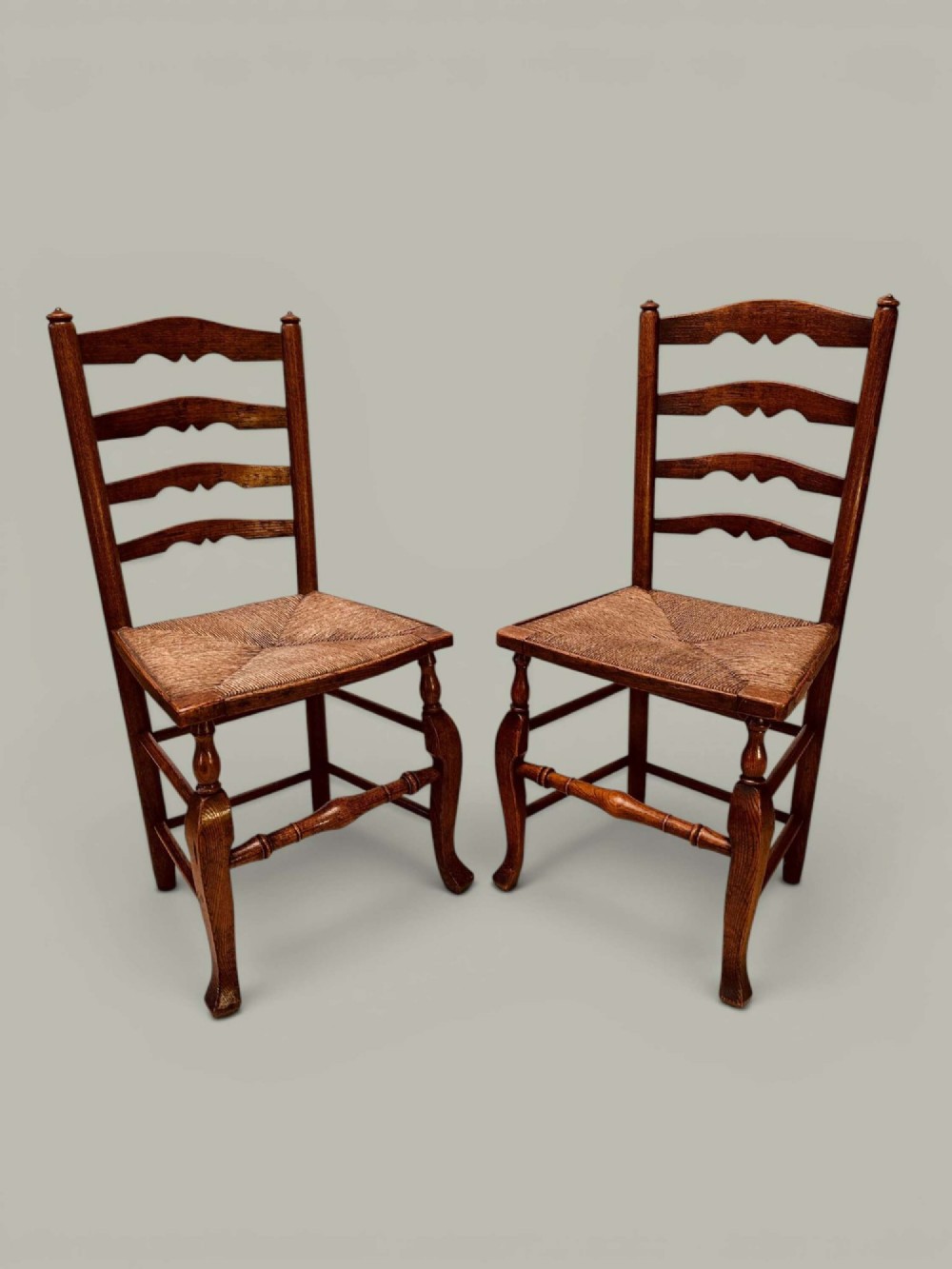 A pair of antique ash ladder back side chairs featuring shaped back rungs, woven rush seats, cabriole front legs, and turned stretchers.