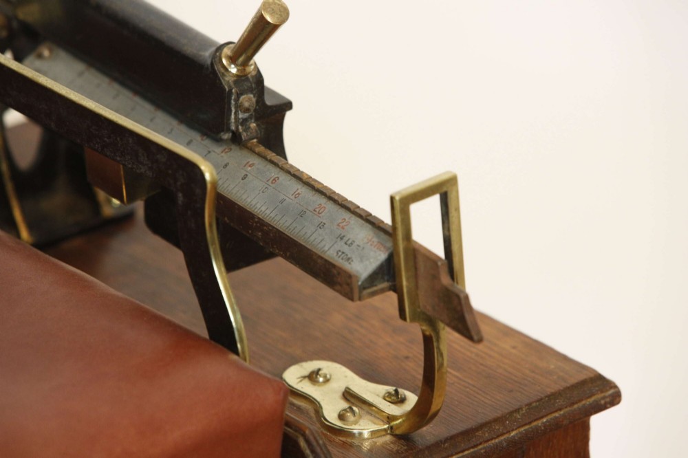 An Edwardian oak jockey scale featuring a padded brown leather seat with brass studs, brass side handles, and an integrated metal balance beam weighing mechanism. View VI
