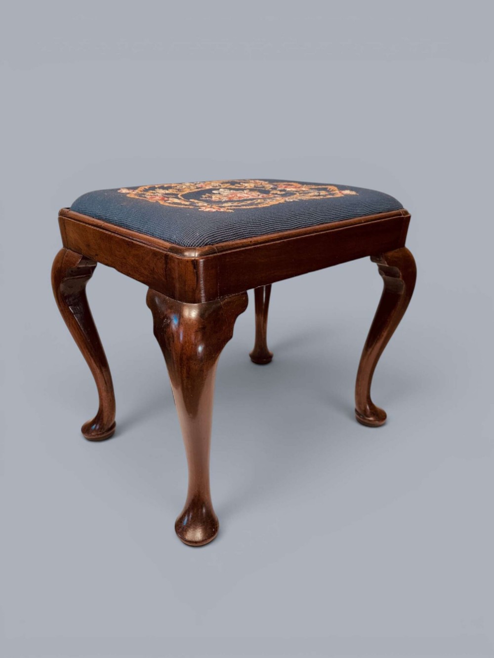 A rectangular red walnut stool with carved cabriole legs, pad feet, and a navy blue needlepoint upholstered seat featuring a central floral wreath motif. View III