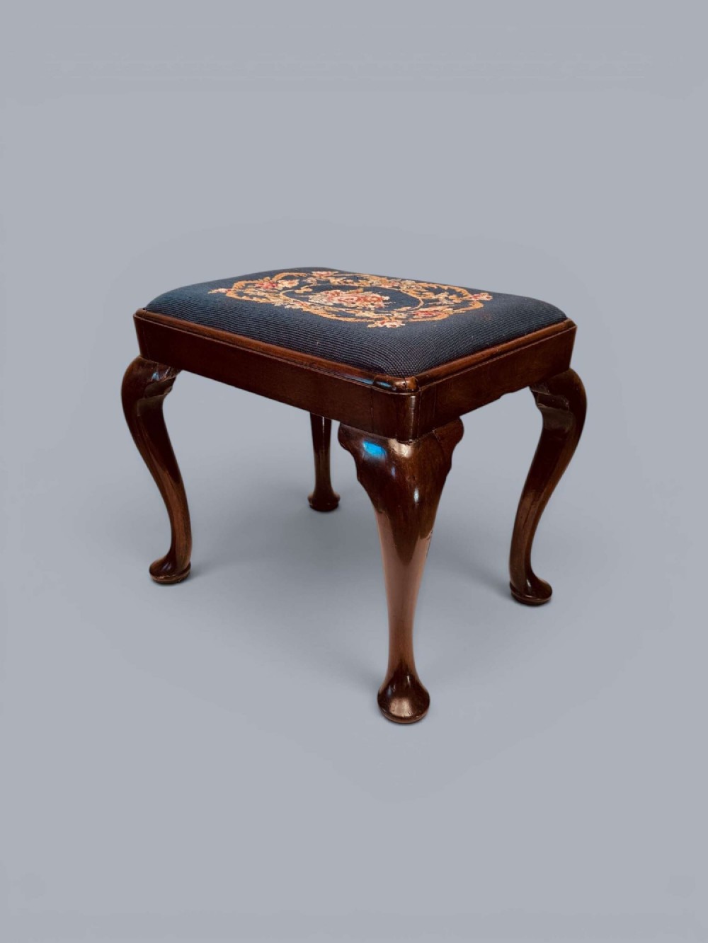 A rectangular red walnut stool with carved cabriole legs, pad feet, and a navy blue needlepoint upholstered seat featuring a central floral wreath motif. View IV