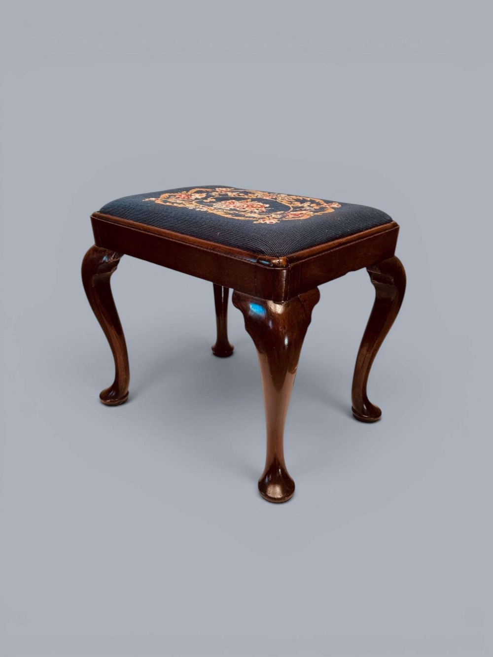 A rectangular red walnut stool with carved cabriole legs, pad feet, and a navy blue needlepoint upholstered seat featuring a central floral wreath motif. View V