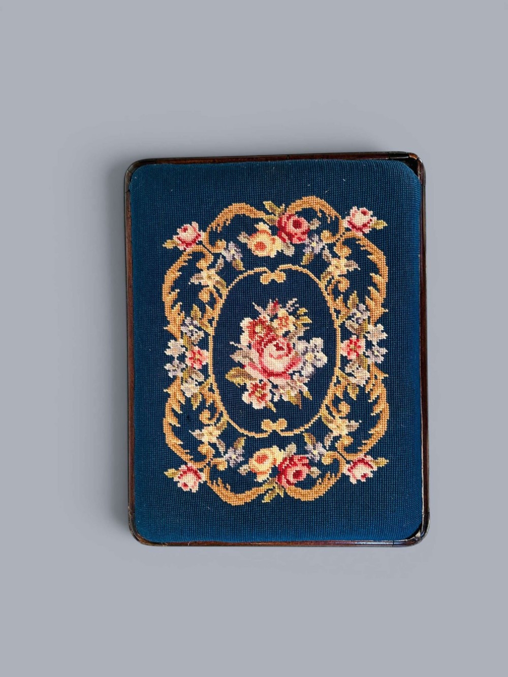 A rectangular red walnut stool with carved cabriole legs, pad feet, and a navy blue needlepoint upholstered seat featuring a central floral wreath motif. View X
