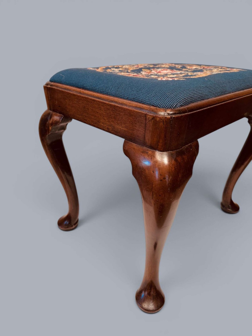 A rectangular red walnut stool with carved cabriole legs, pad feet, and a navy blue needlepoint upholstered seat featuring a central floral wreath motif. View XII