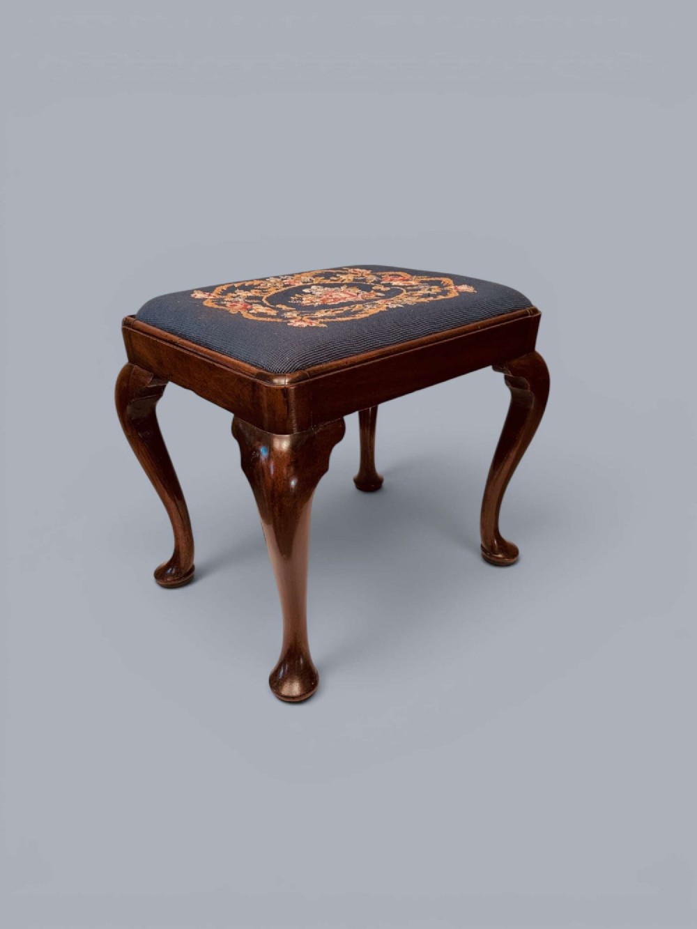 A rectangular red walnut stool with carved cabriole legs, pad feet, and a navy blue needlepoint upholstered seat featuring a central floral wreath motif. View XIII