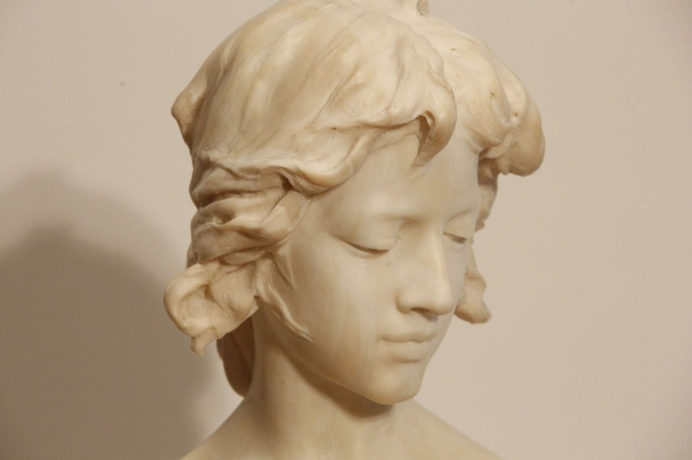 An early twentieth-century carved Carrara marble bust depicting a young female subject with a carved floral corsage, mounted on a wooden pedestal base. View IV