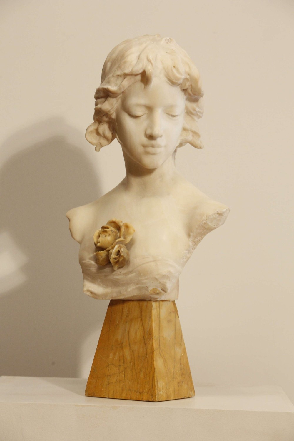 An early twentieth-century carved Carrara marble bust depicting a young female subject with a carved floral corsage, mounted on a wooden pedestal base. View X