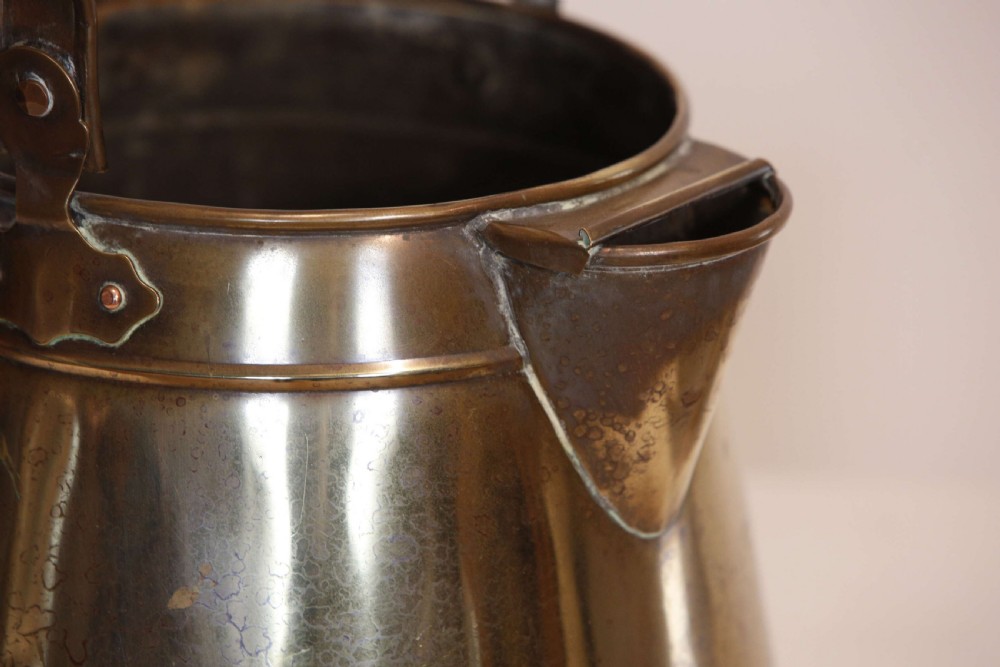 An antique brass hot water can featuring a tapered cylindrical body, broad overhead swinging handle, rear side handle, and riveted construction. View VI