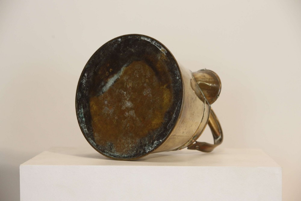 An antique brass hot water can featuring a tapered cylindrical body, broad overhead swinging handle, rear side handle, and riveted construction. View X