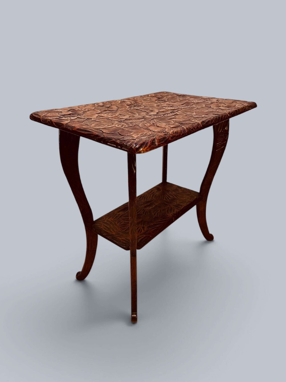 A two-tier carved hardwood side table with splayed legs and relief-carved leaf patterns across the tabletop and lower shelf.