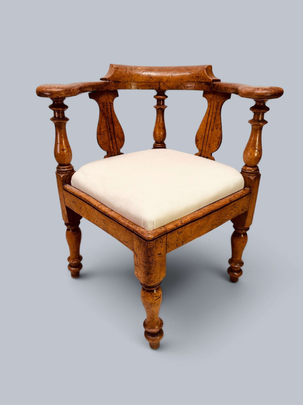 A Victorian corner chair crafted from burr elm featuring turned upright supports, shaped back splats, a curved top rail, and a cream upholstered drop-in seat.