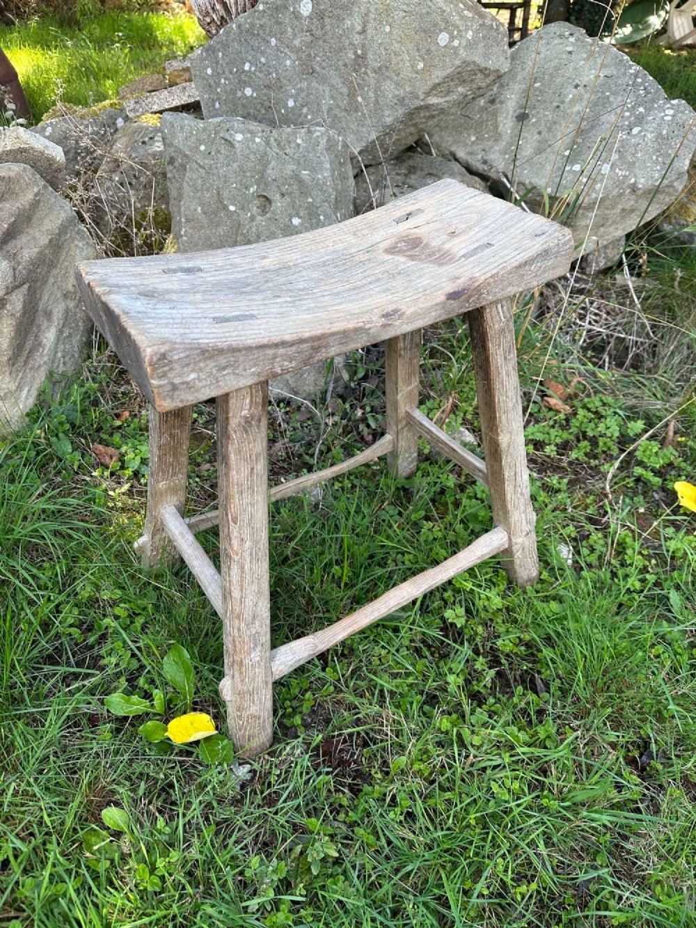 An antique weathered grey Chinese elm stool featuring a curved concave wooden seat, splayed legs, and exposed tenon joinery.