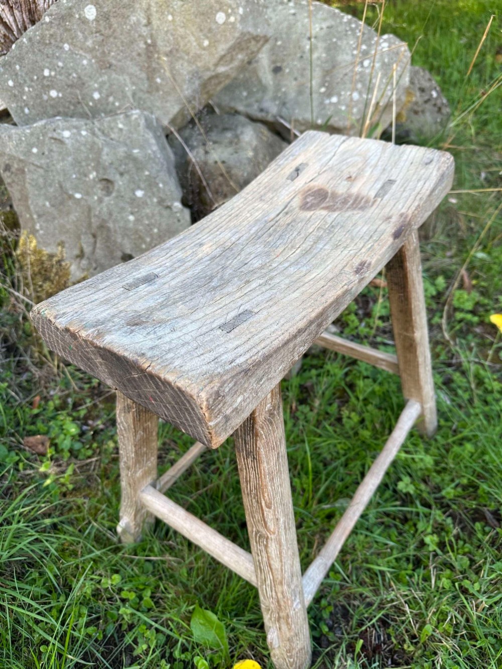 An antique weathered grey Chinese elm stool featuring a curved concave wooden seat, splayed legs, and exposed tenon joinery. View VI