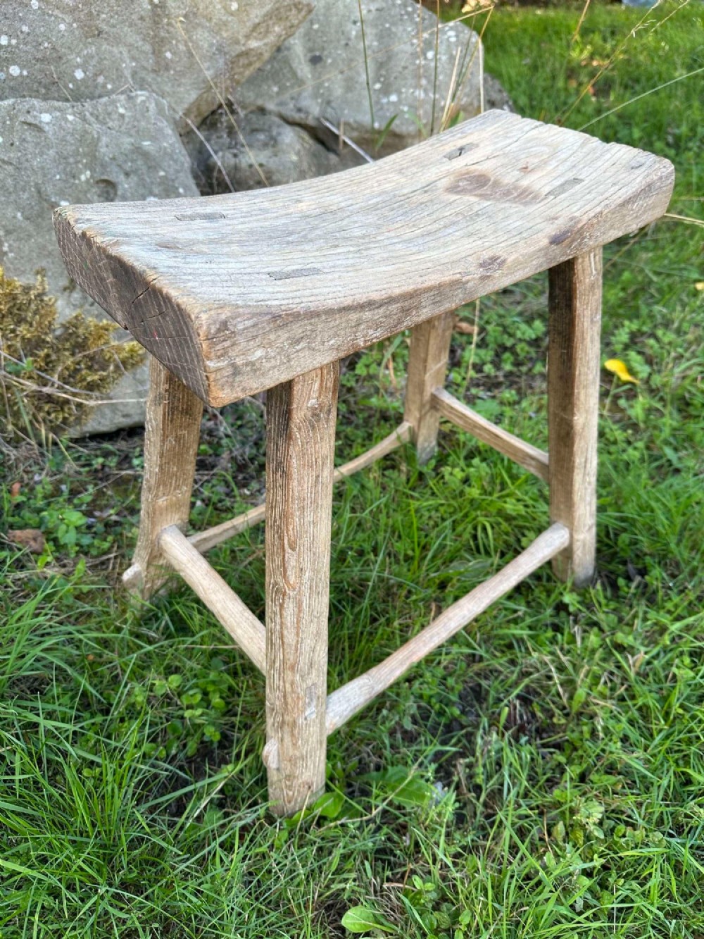 An antique weathered grey Chinese elm stool featuring a curved concave wooden seat, splayed legs, and exposed tenon joinery. View IX