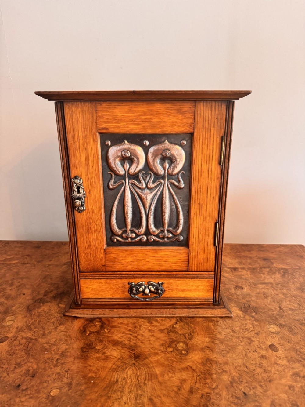 An Art Nouveau golden oak smokers cabinet featuring a single door inset with an embossed copper panel above a lower drawer.