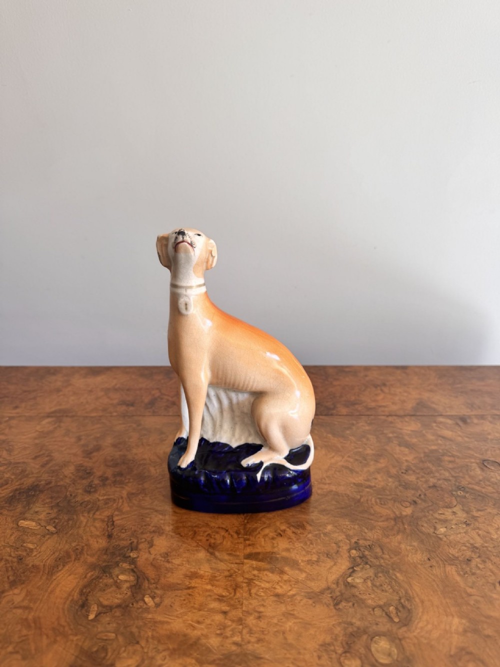 A Victorian Staffordshire ceramic figurine depicting a seated tan and white greyhound set upon a cobalt blue base.