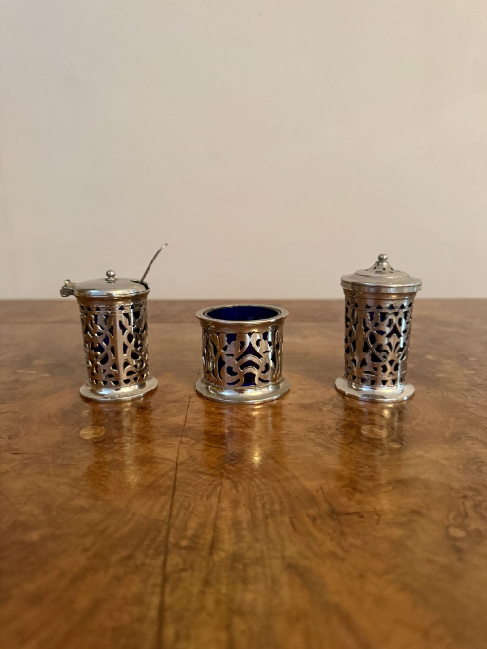 A three-piece Edwardian silver cruet set featuring openwork scroll patterns and blue glass insert liners, comprising a mustard pot with spoon, open salt cellar, and pepper shaker.
