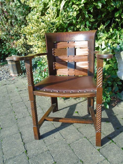 fig 1. An Arts and Crafts solid oak armchair with carved spiral front legs, a brown leather seat secured with studs, and a curved slatted backrest.
