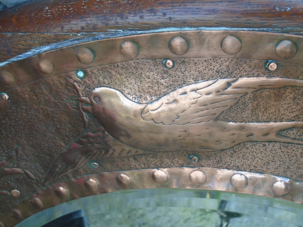 A circular antique Arts and Crafts copper wall mirror featuring hand-hammered repousse decoration of birds, foliage, and urns with a riveted border. View V
