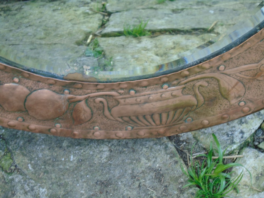 A circular antique Arts and Crafts copper wall mirror featuring hand-hammered repousse decoration of birds, foliage, and urns with a riveted border. View VI