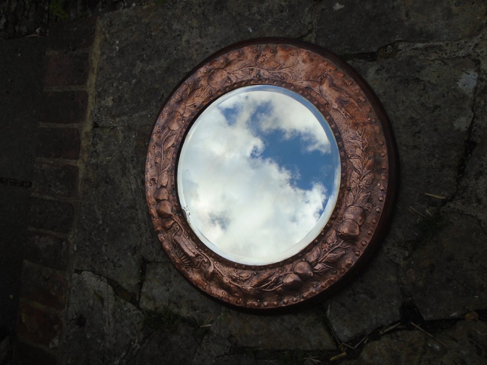 A circular antique Arts and Crafts copper wall mirror featuring hand-hammered repousse decoration of birds, foliage, and urns with a riveted border. View XII