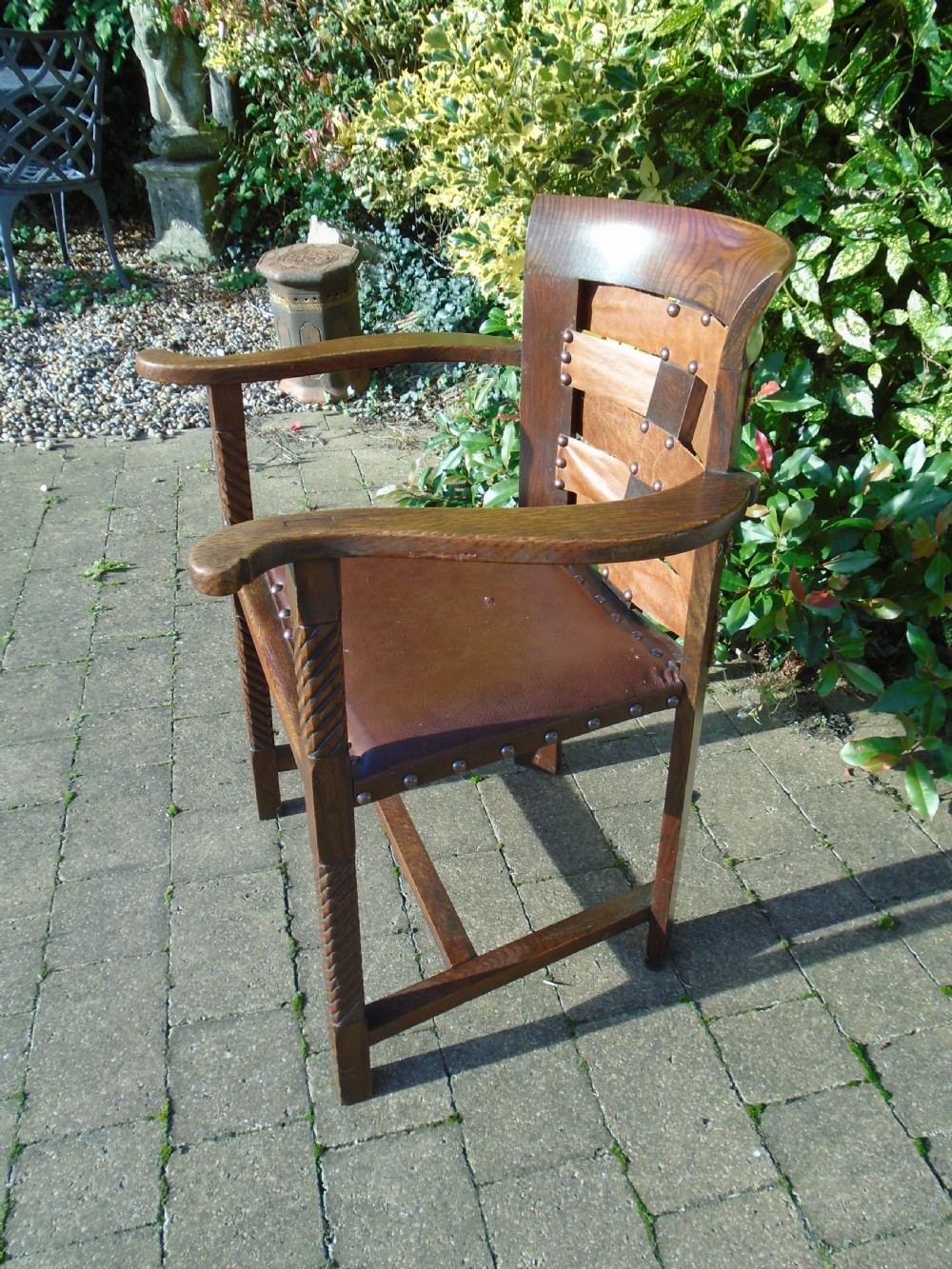 An Arts and Crafts solid oak armchair with carved spiral front legs, a brown leather seat secured with studs, and a curved slatted backrest. View V