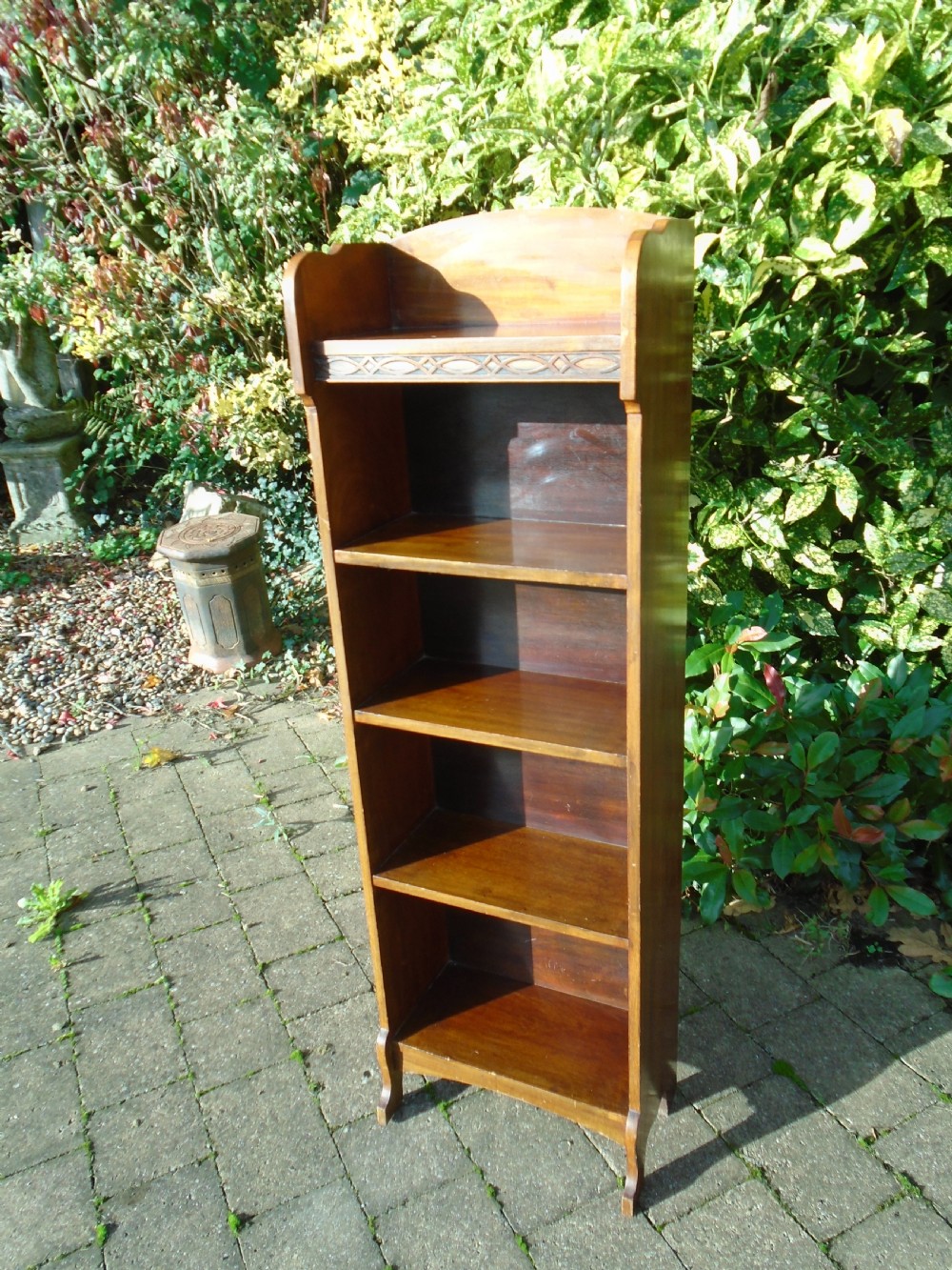 A tall, narrow Arts and Crafts walnut open bookcase featuring four shelves and carved decorative details along the upper frieze.