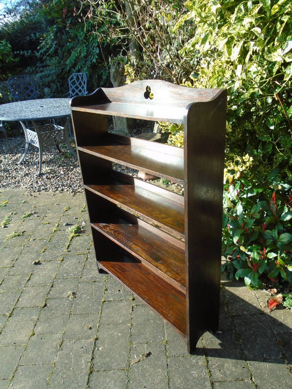 A dark oak Arts and Crafts open bookcase featuring four shelves and a shaped gallery top with a trefoil cutout motif.