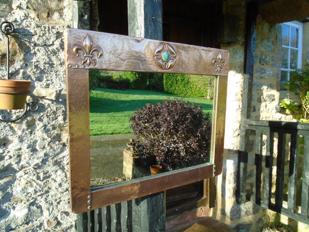 A rectangular hand-hammered copper wall mirror featuring raised fleur-de-lis motifs in the top corners and a central green glass cabochon.