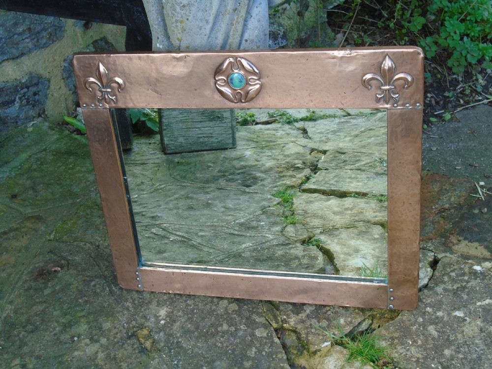A rectangular hand-hammered copper wall mirror featuring raised fleur-de-lis motifs in the top corners and a central green glass cabochon. View II