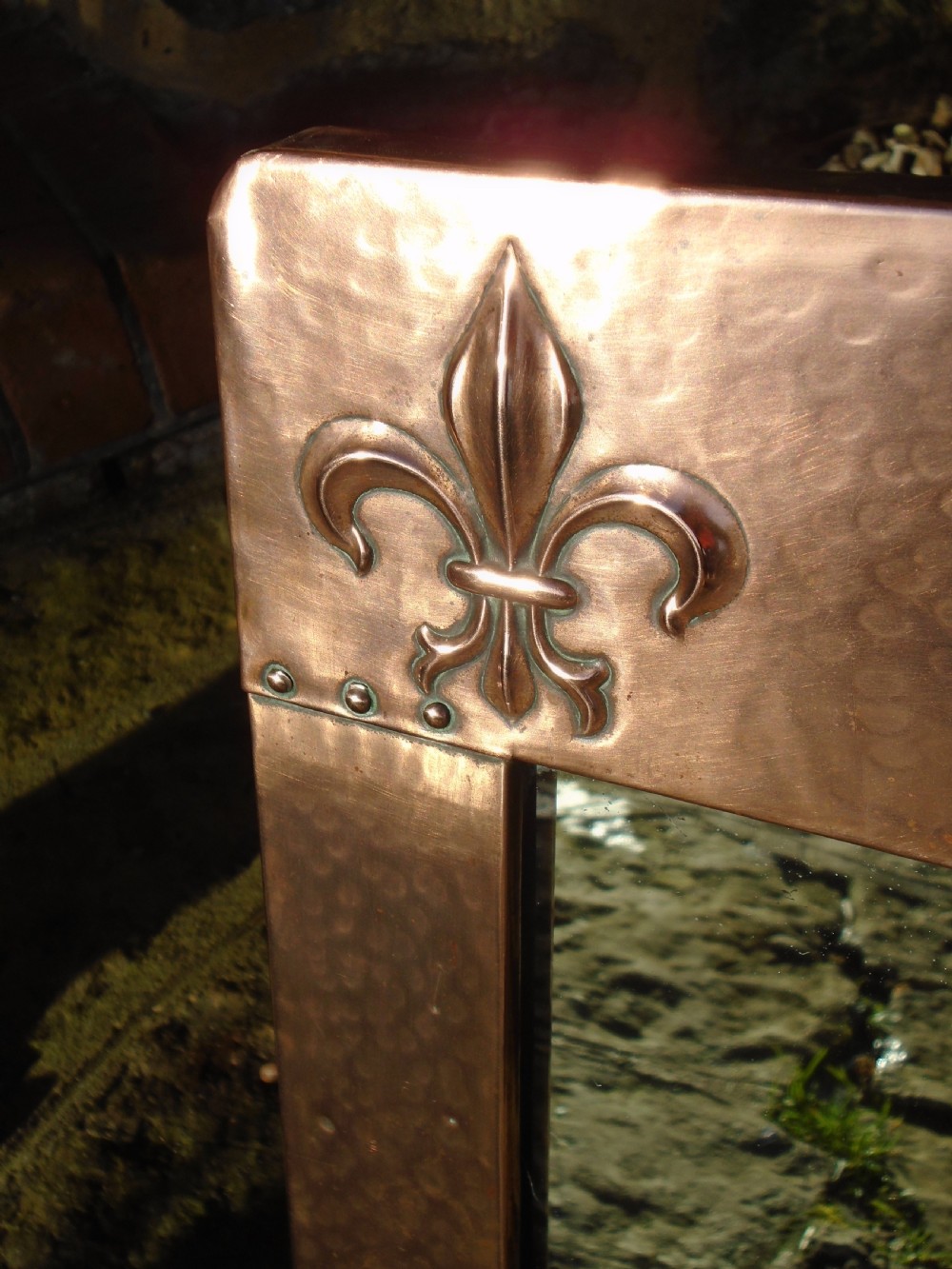 A rectangular hand-hammered copper wall mirror featuring raised fleur-de-lis motifs in the top corners and a central green glass cabochon. View III