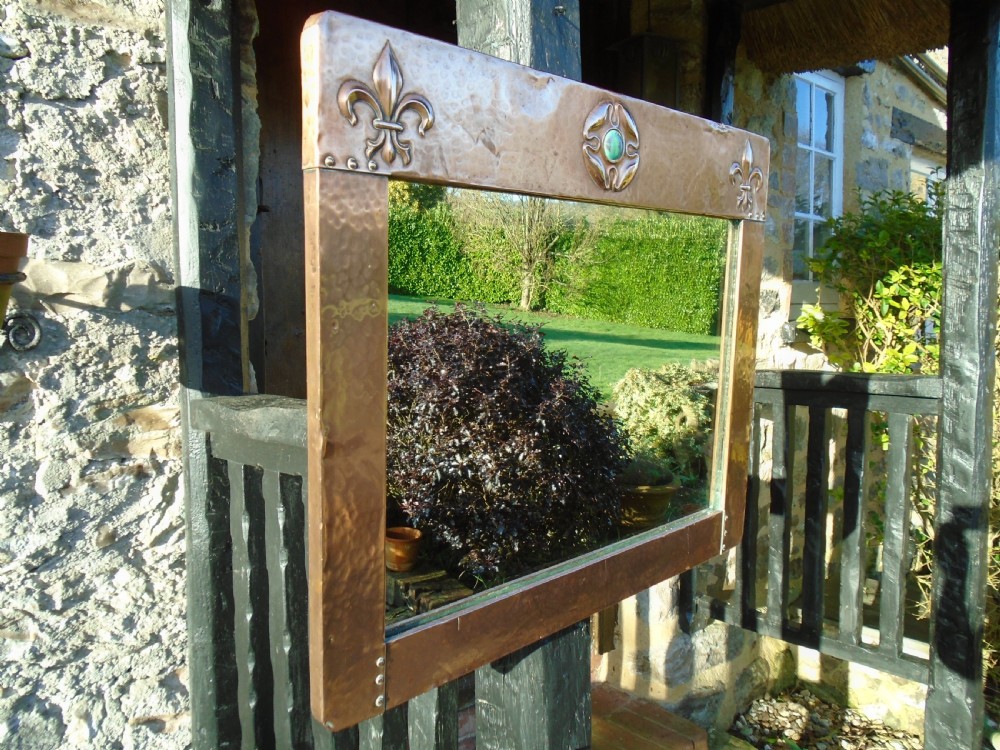 A rectangular hand-hammered copper wall mirror featuring raised fleur-de-lis motifs in the top corners and a central green glass cabochon. View IV