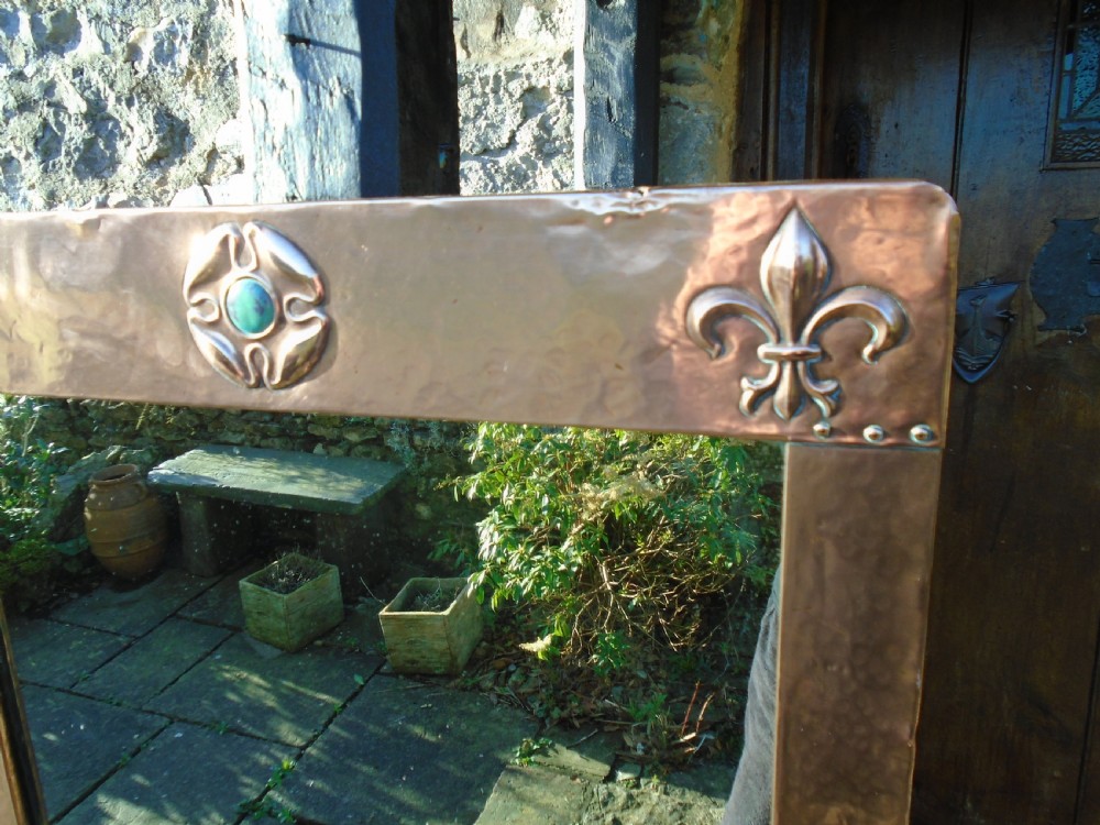A rectangular hand-hammered copper wall mirror featuring raised fleur-de-lis motifs in the top corners and a central green glass cabochon. View V
