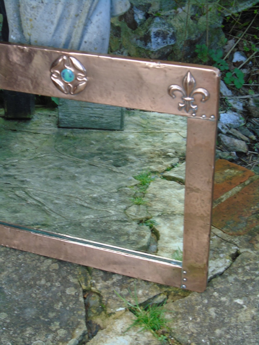 A rectangular hand-hammered copper wall mirror featuring raised fleur-de-lis motifs in the top corners and a central green glass cabochon. View VI