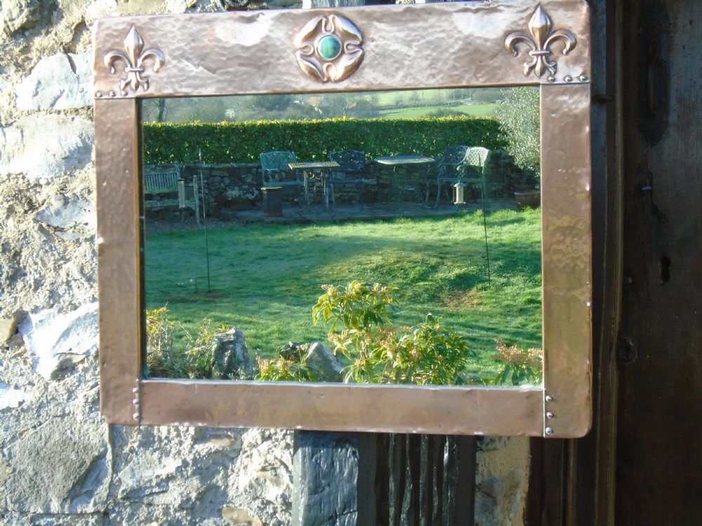 A rectangular hand-hammered copper wall mirror featuring raised fleur-de-lis motifs in the top corners and a central green glass cabochon. View VII